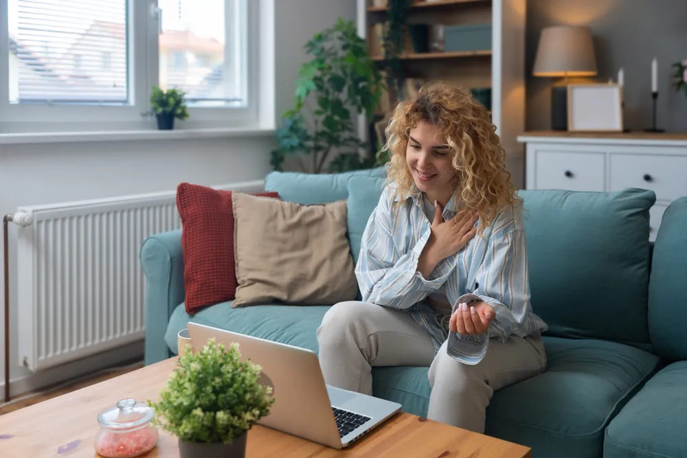 A woman sitting on her couch at home, smiling as she talks with someone on her laptop